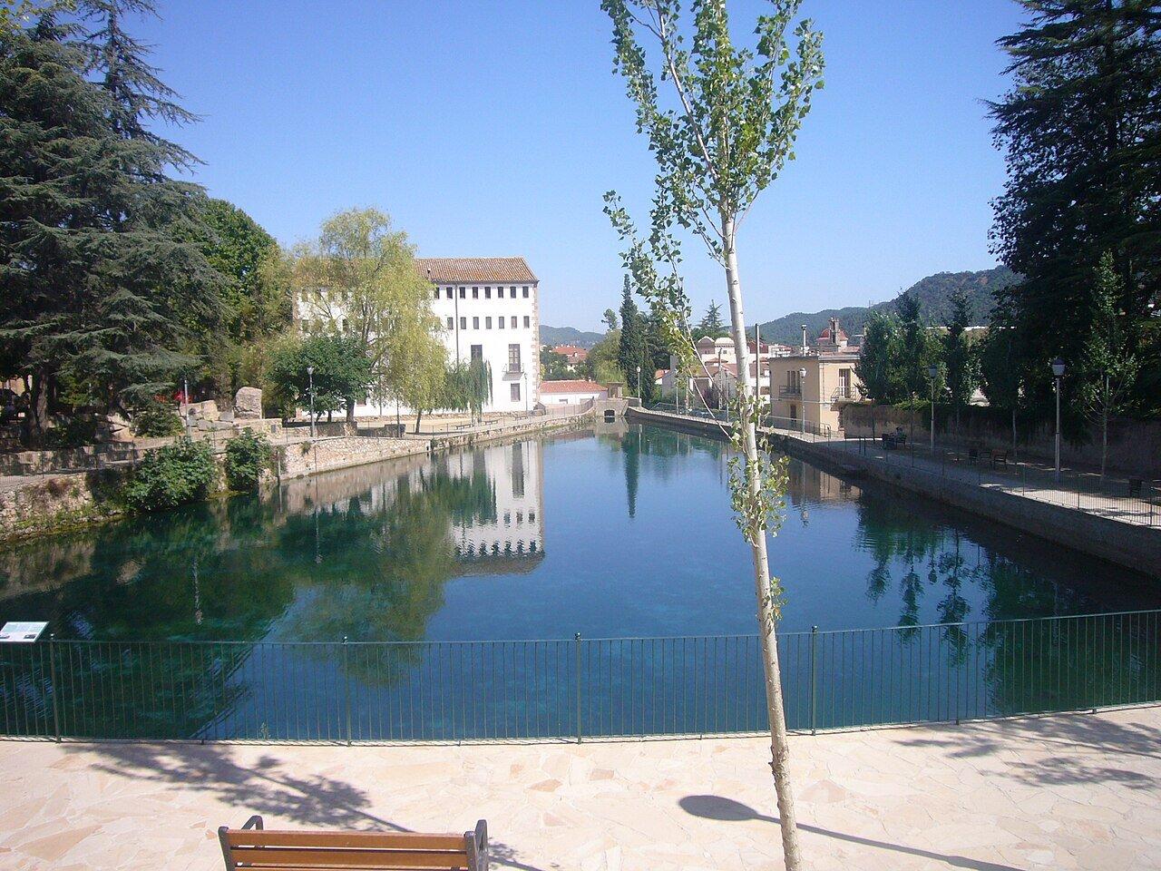The Bassa de Capellades reflecting the paper mill and trees beside a waterside path